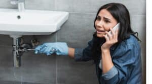 Stressed girl repairing pipe with wrench and talking on smartphone